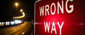 Close-up of a red 'WRONG WAY' road sign at night, with blurred highway lights in the background.